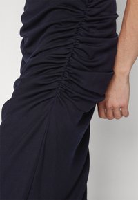 Abito blu navy con un design arricciato lungo il lato e una texture liscia. Il tessuto è morbido, aderente e cade elegantemente.