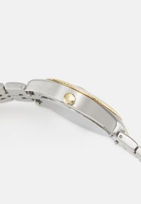 Silverfärgad metallklocka med en guldkant. Har en texturerad krona och en polerad yta. Armbandet består av länkade metalldelar.
