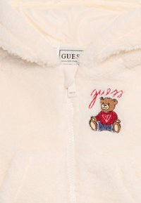 Giacca in pile color crema con zip, orsetto ricamato seduto che indossa un maglione rosso, e testo "Guess" sopra.