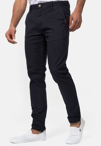 Chinos de algodón azul marino con un corte ajustado, cierre de botón, bolsillos laterales y un pequeño parche de marca en la parte frontal. Combinados con zapatos blancos tipo slip-on.