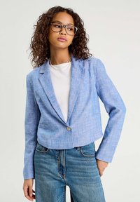 Blazer bleu cintré avec une finition texturée, fermeture à un bouton et revers crantés, associé à un jean en denim taille haute.