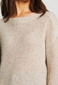 Close-up of a woman wearing a beige knitted sweater with a ribbed round neckline and speckled yarn details.