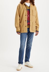 Chaqueta de mezcla de lana color marrón con dos bolsillos frontales, combinada con un suéter de rombos en color burdeos, azul y amarillo, jeans azules y zapatillas blancas.