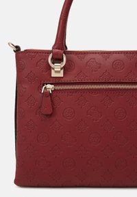 Punainen synteettinen käsilaukku, jossa on embossattu geometrinen kuvio, kultasävyiset varusteet, vetoketjullinen tasku ja sileä, teksturoitu pinta.