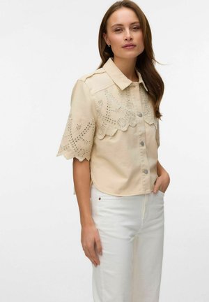 Femme portant une chemise beige à manches courtes avec des motifs floraux découpés et un pantalon taille haute blanc sur un fond blanc.