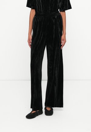 Zwarte velours jumpsuit met korte mouwen, wijde broekspijpen, elastische tailleband, met een zachte textuur en verticale streepjespatroon. Zwarte platte schoenen.