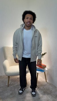 Beige zip-up hoodie over a white t-shirt, black jeans, and black sneakers with white accents. Soft textured chair and books in background.