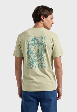 Hombre con camiseta beige con dibujo de línea azul de un pueblo costero, edificios, palmera, farola, mesa con vaso y pez en la parte de atrás.