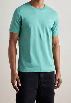 Mężczyzna w jednokolorowym, turkusowym t-shircie z krótkim rękawem i okrągłym dekoltem oraz czarnych spodniach, stojący z rozluźnionymi rękami wzdłuż ciała na jednolitym tle.