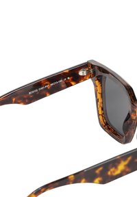 Okulary przeciwsłoneczne w musztardowym kolorze z ciemnymi soczewkami, szerokimi kwadratowymi oprawkami i błyszczącym wykończeniem. Szczegóły obejmują metalowe akcenty i wygrawerowane informacje o modelu.