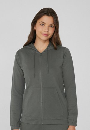Junge Frau mit langen braunen Haaren trägt einen offenen, dunkelgrauen Hoodie mit vorderen Taschen und steht vor einem einfarbigen hellgrauen Hintergrund.