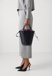 Black leather bucket bag with drawstring closure, embossed "COCO" logo, paired with a checked suit and black high-heeled shoes.