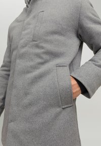 Grå uldfrakke med en glat tekstur, der har en enkelt brystlomme og et strømlinet design. Stoffet er varmt og tykt.