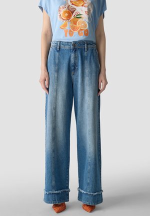 Person trägt hellblaues T-Shirt mit orangefarbenem Fruchtmotiv und Text, weit geschnittene blaue Jeans mit ausgefransten Säumen und orangefarbene gewebte Schuhe.