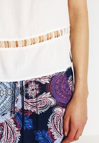 Top bianco testurizzato con dettagli a taglio orizzontale, abbinato a una parte inferiore con motivo paisley blu e rosso. Focalizzati sul materiale e sul design colorato.