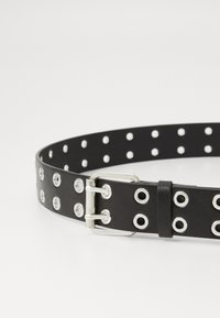 Ceinture en cuir noir avec des œillets en métal argenté et une boucle en métal. Présente des trous espacés de manière égale pour un ajustement réglable. Texture lisse.