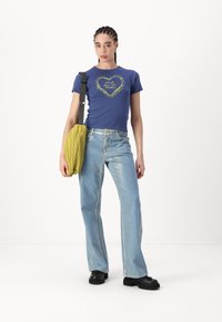 Zila kokvilnas t-kreklu ar sirds grafiku zaļā krāsā, apvienota ar gaišziliem platā krokā esošiem džinsiem un saburzītu dzeltenu crossbody somu. Melni apavi.