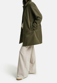 Osoba oblečená v olivově zeleném zapínac�ím kabátu, béžových širokých kalhotách a bílých teniskách, stojící s jednou rukou v kapse kabátu.