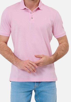 Man draagt een lichtroze poloshirt met korte mouwen en twee knopen en lichtblauwe spijkerbroek, handen zachtjes voor zich gevouwen.