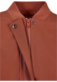 Jacket v rustovej farbe s vysokým golierom, zipsom na prednej strane, kovovým zacvakávacím uzáverom a hladkým textúrovaným materiálom.
