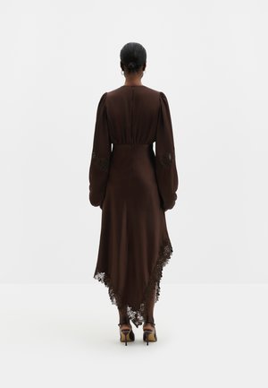 Femme aux cheveux foncés en chignon bas, portant une longue robe marron foncé avec des détails en dentelle et des manches longues, debout de dos sur un fond blanc.