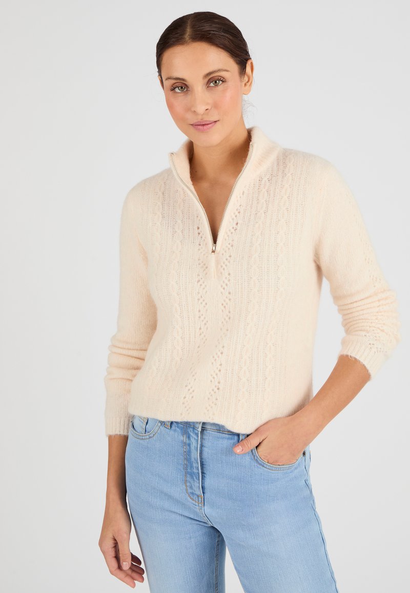 Suéter de punto rosa claro con cuello de cremallera, que presenta un patrón texturizado y puños acanalados. Combinado con jeans azules, mostrando un look casual.
