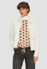 Witte fleece jas met een kraag, twee voorzakken en zilveren drukknoopjes, gedragen over een geometrisch patroon top en donkere broeken.