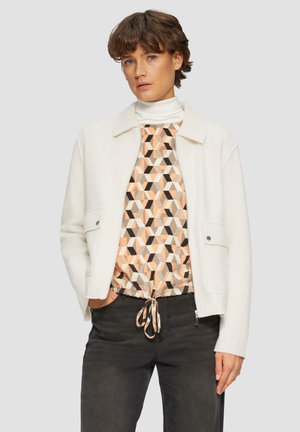 White fleece jacket with a collar, two front pockets, and silver snap buttons, worn over a geometric patterned top and dark pants.