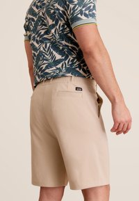Beige shorts met een elastische tailleband, een achterzak en een label. Het patroon op het shirt toont groene bladeren op een crèmekleurige achtergrond met ribbelmanchetten.
