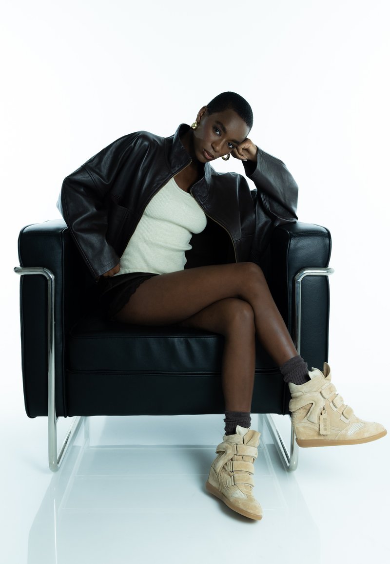 Veste surdimensionnée en cuir marron, débardeur côtelé crème, shorts noirs, baskets compensées beige à trois brides, sur une chaise noire sur un fond blanc.