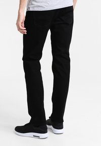 Mężczyzna w wąskich czarnych dżinsach, jasnoszarej koszulce i czarnych sneakersach z białymi podeszwami, stoi na jednolitym tle.