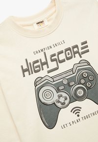 Kreemi värvi T-särk, millel on trükitud mängukontrolleri graafika, koos julge tüpograafiaga "HIGH SCORE" ja "LET'S PLAY TOGETHER".