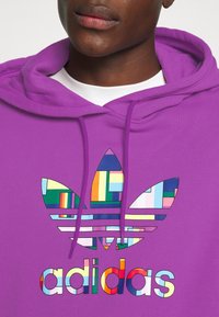 Lila huvtröja med en färgglad Adidas-logotyp med geometriska mönster och ränder. Texten "adidas" i flerfärgad skrift nedanför.