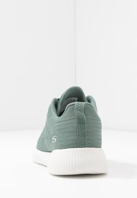 Grön stickad tygsneaker med vit sula, sett bakifrån, med en liten sidologga och dragflik på hälen.