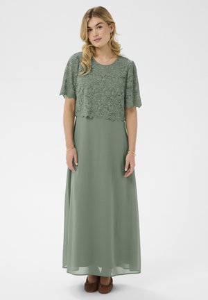 Kobieta w zieloną suknię maxi w kolorze szałwii z koronkową, krótką narzutką, stojąca na jasnym jednolitym tle.