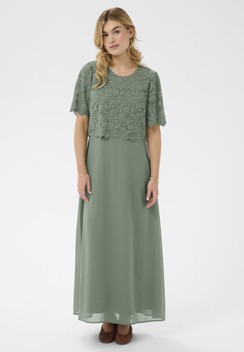 Femme portant une robe maxi vert sauge avec une superposition en dentelle à manches courtes, debout devant un fond clair uni.