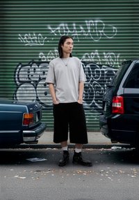 T-shirt cinzenta oversize, calções pretos até ao joelho e botas pretas até ao tornozelo com meias brancas. Em pé entre dois carros contra um fundo de graffiti.