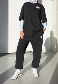 Schwarzes Baumwoll-T-Shirt mit einem Logo-Patch, kombiniert mit passenden schwarzen Hosen mit Taschen. Weiße Sneakers runden das Outfit ab.