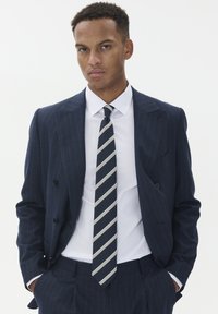 Jūržalių juostelių kostiumas su dvigubos krūtinės švarku, balta marškiniai ir juostuota kaklaraiščiu, kuriame dominuoja jūržalių, pilkų ir sidabrinių spalvų.