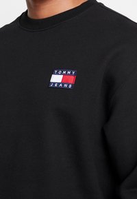 Felpa nera con colletto a costine e un logo ricamato rettangolare in blu, bianco e rosso con il testo "TOMMY JEANS" sottostante.