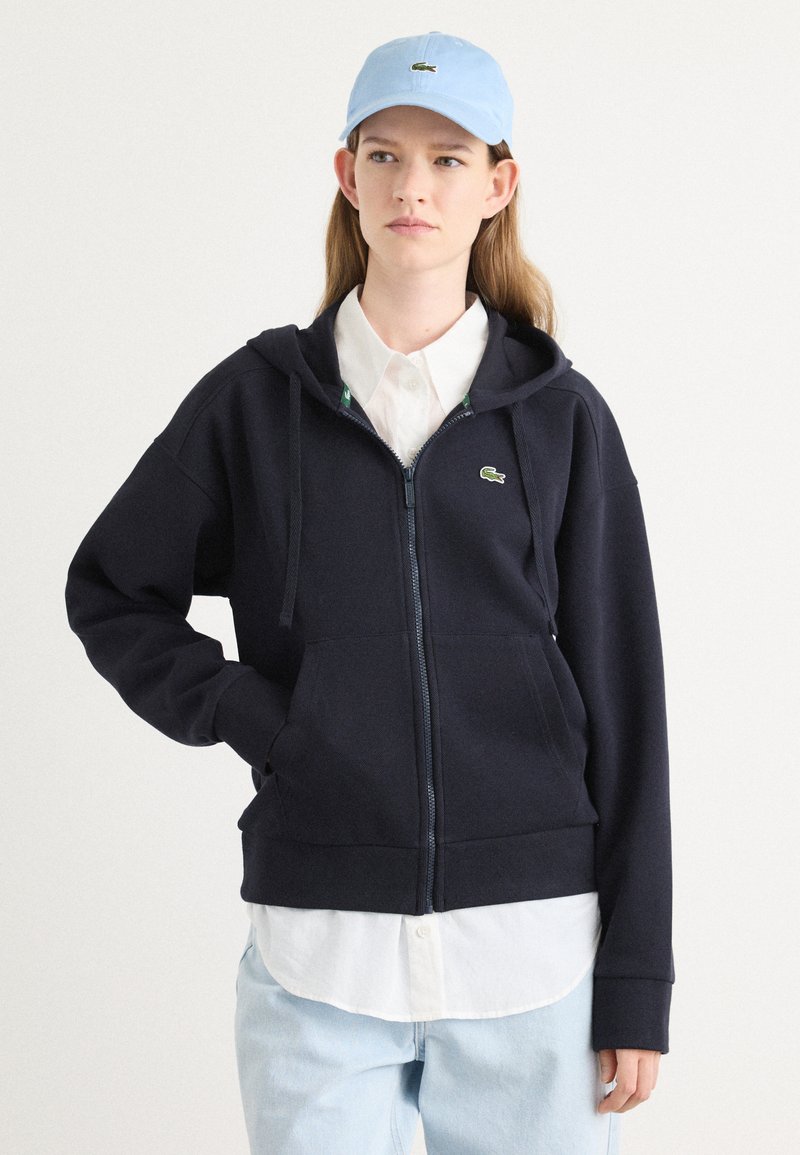 Sudadera con cremallera en color marino, fabricada en un tejido suave, con un bolsillo canguro y capucha ajustable. Combinada con una camisa blanca de botones y una gorra de color azul claro.