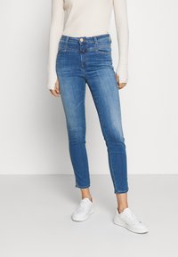 Úzké modré džíny s vysokým pasem, vyrobené z denimu. Džíny mají klasický design s pěti kapsami a jemným vyblednutím, kombinované s bílými teniskami.