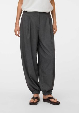 Personne portant un pantalon ample gris foncé avec des chevilles élastiques et des sandales à plateforme noires, debout devant un fond uni.