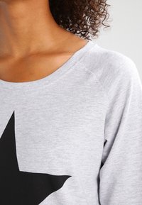 Sudadera gris con cuello redondeado y gráfico de estrella negra. Textura suave, corte ligeramente holgado y diseño de manga raglán.