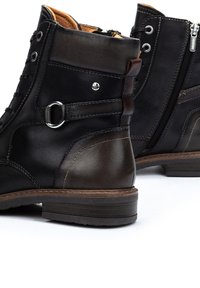 Bottes en cuir noir avec un accent marron. Les caractéristiques comprennent un anneau en métal circulaire, une fermeture éclair sur le côté et des détails de couture contrastants.