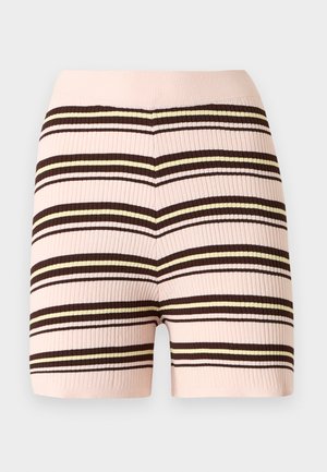 Kvinne står med hånden på hoften, iført ermeløs stripete topp, rosa og brune stripete shorts, og svarte flate sko med spenne.