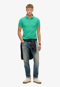 Polo verde de corte ajustado, jeans azules y una camisa de cuadros atada a la cintura. Zapatillas blancas con suela marrón.