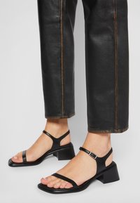 Pieds portant des sandales en cuir noir à bouts carrés avec des talons carrés bas et des brides à la cheville, associées à un pantalon en cuir noir raccourci.