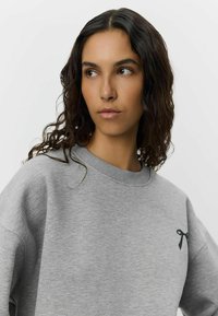 Grey sweatshirt made of soft fabric, featuring a crew neck and a black ribbon detail on the left chest. Relaxed fit with dropped shoulders.