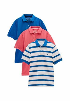 Mand iført en hvid og blåstribet polotrøje og lyserøde shorts stående mod en ensfarvet beige baggrund.